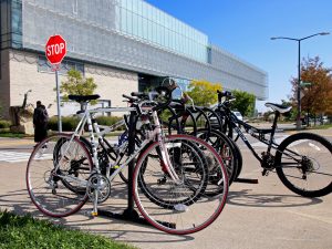 Several bicycles are locked on a tall bicycle rack near Brock University’s Cairns Complex. In the background is a stop sign and a person crossing the street.