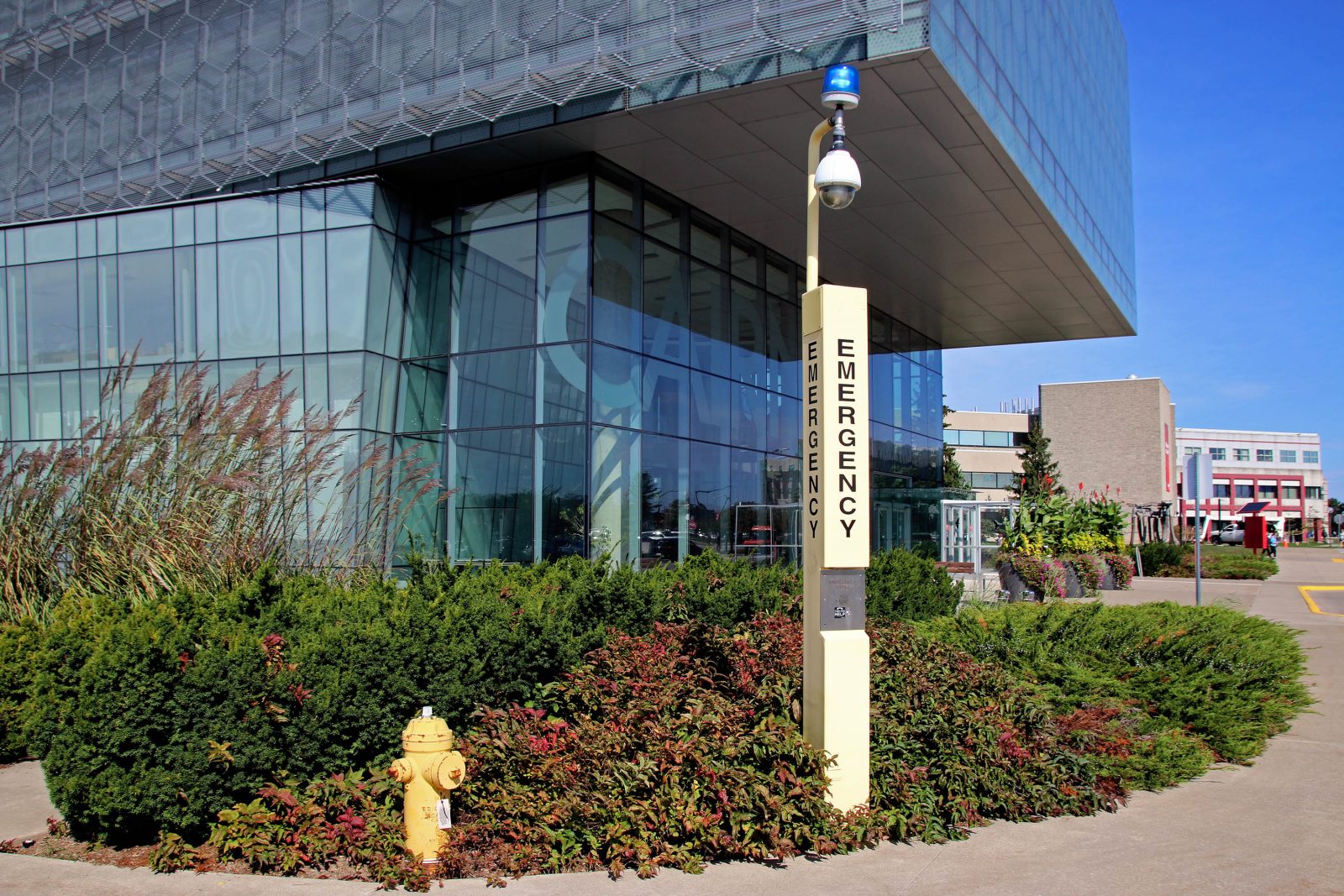 A large emergency phone — a tall yellow pole with “EMERGENCY” written on it and a security camera and blue light attached to the top — is seen in front of a large glass building with “CAIRNS” written on it. A fire hydrant is next to the phone, and other buildings can be seen in the background.