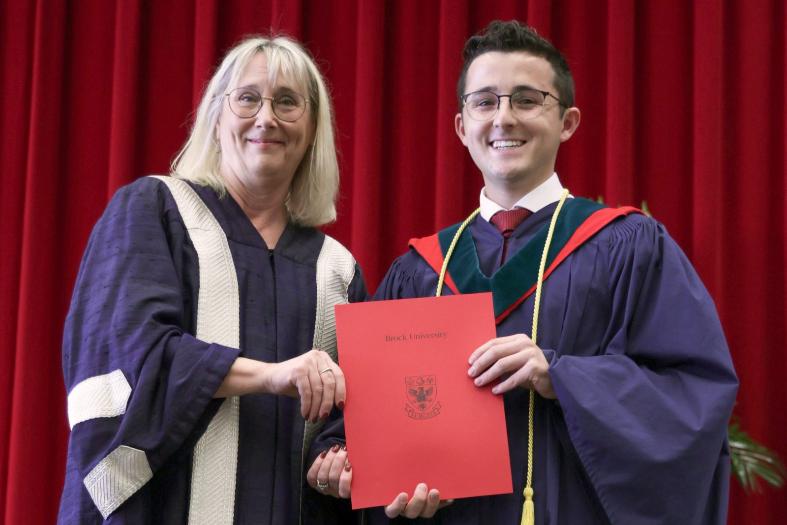 A woman and a man in graduation robes hold a red certificate between them.