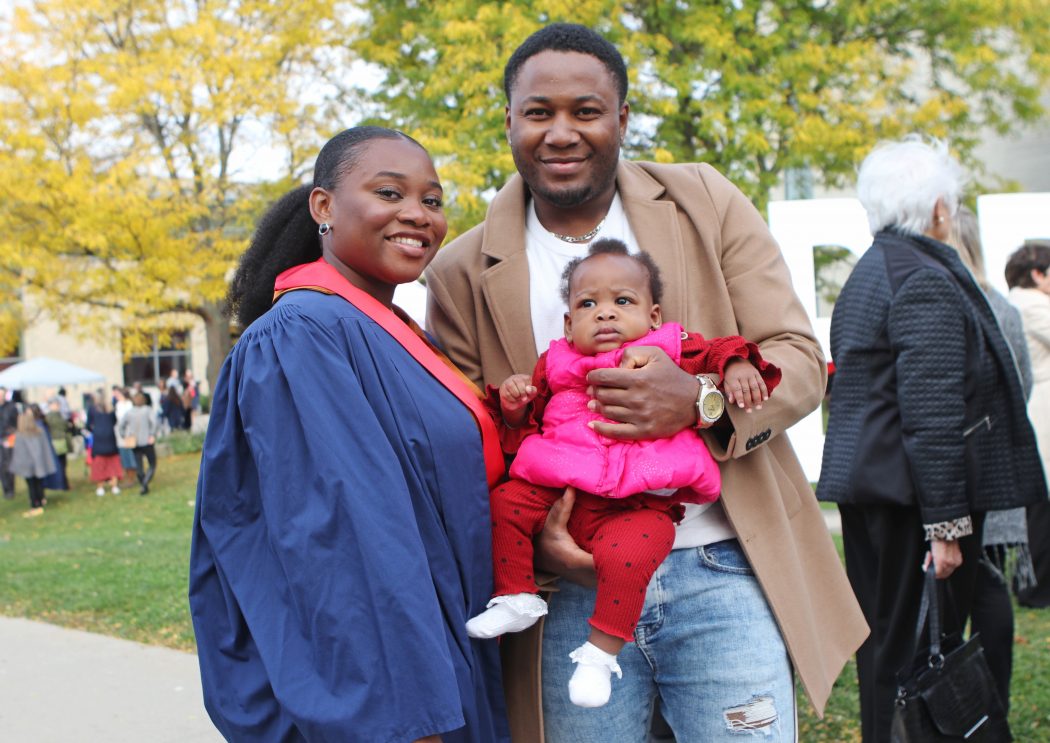 A woman in a blue graduation gown stands beside a man holding a baby.