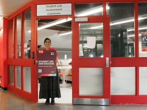 A student in a red shirt stands in a doorway holding a purple sign.