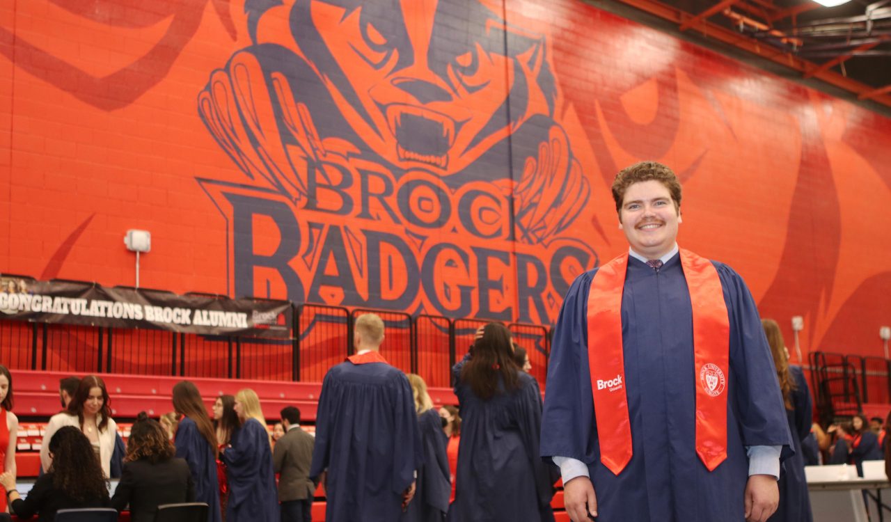 A man in a blue robe and red sash stands in front of a large red brick wall that says "Brock Badgers" on it.