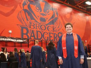 A man in a blue robe and red sash stands in front of a large red brick wall that says "Brock Badgers" on it.