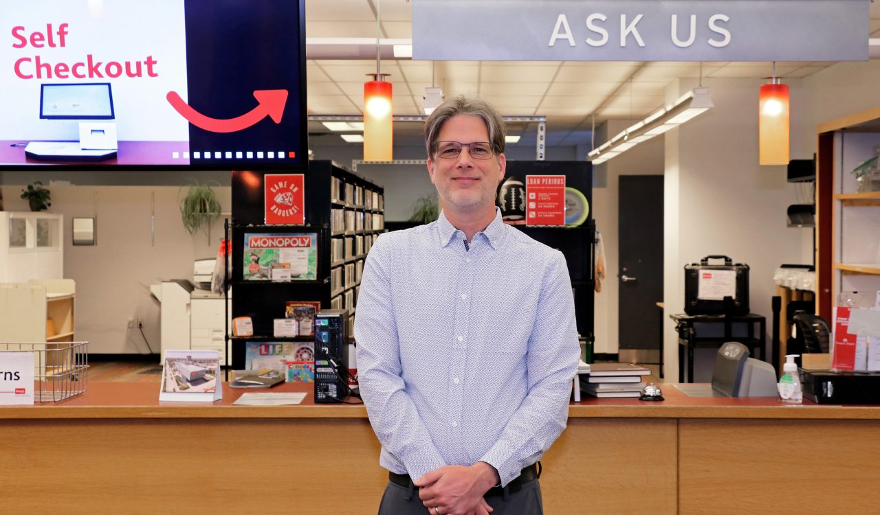 A man stands in front of a desk area in a university library. A sign above his head reads “Ask Us” and a digital screen displays instructions for where to use the self-checkout machines.
