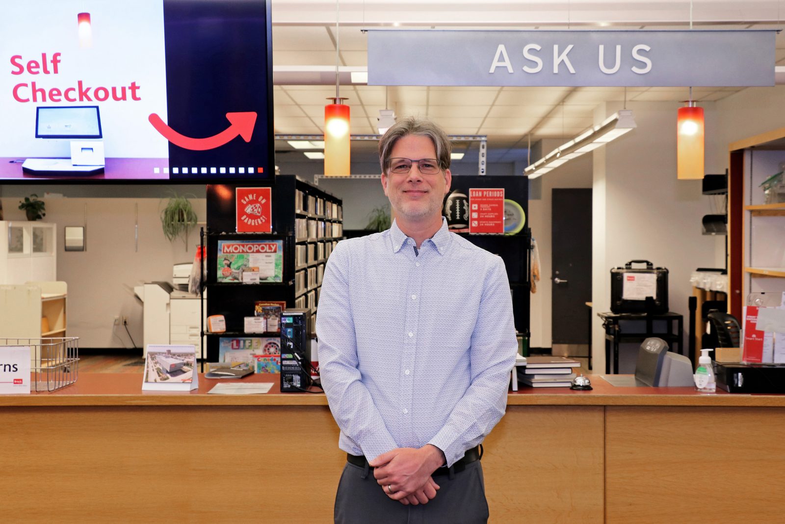 A man stands in front of a desk area in a university library. A sign above his head reads “Ask Us” and a digital screen displays instructions for where to use the self-checkout machines.