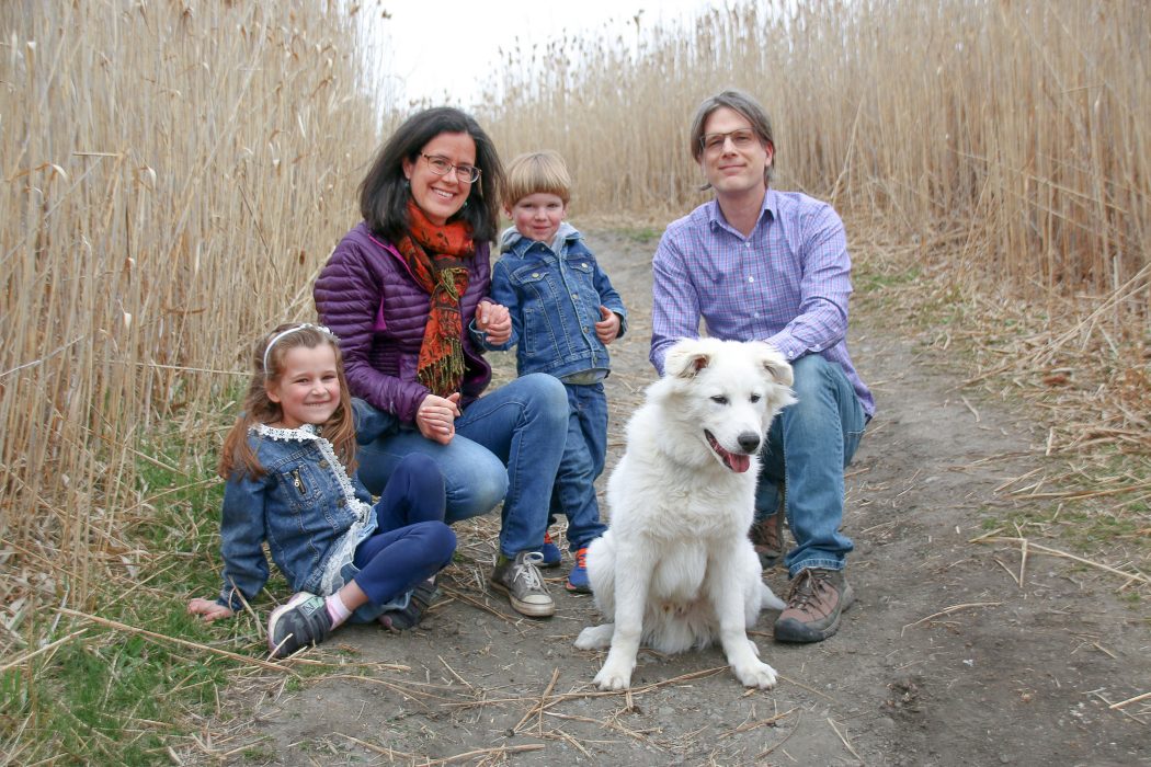 Two adults and two children kneel on a dirt path flanked by dried, tall grasses. Their pet, a tall white dog, stands beside them.