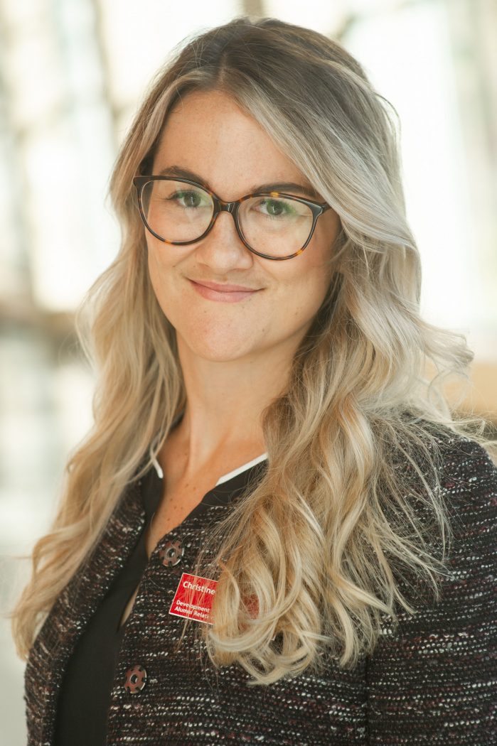 A woman in a brown and black blazer stands in front of windows with sun shining through them