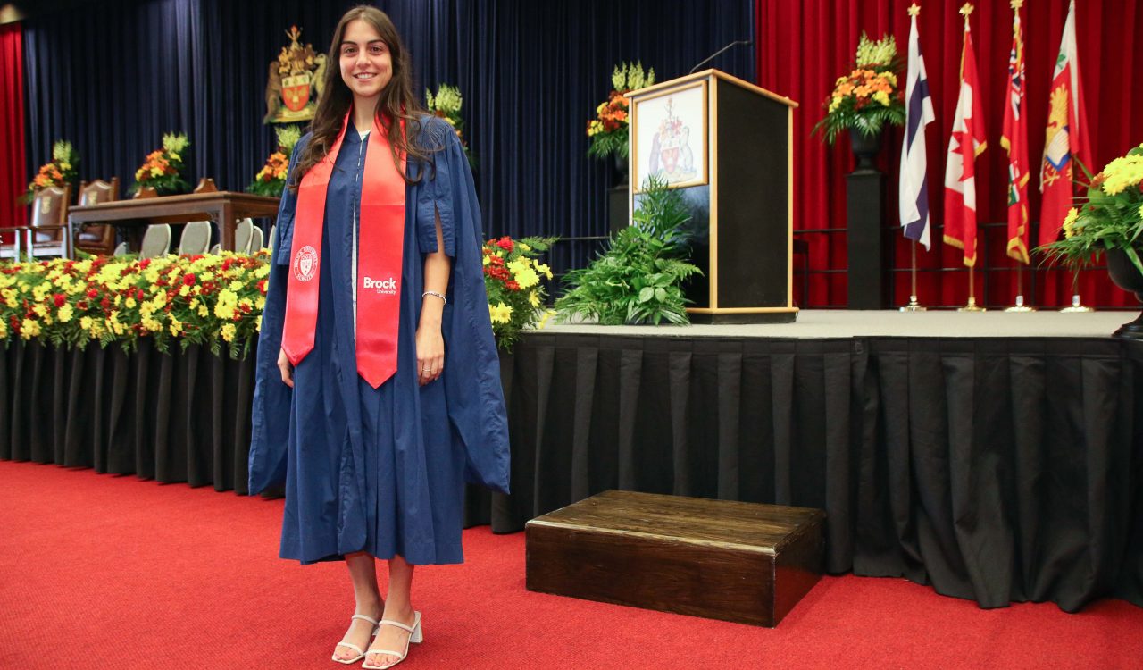 A woman in a blue convocation robe and red sash stands in front of a stage decorated with flowers and a lectern.