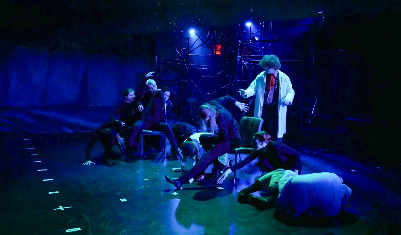 A dark stage with blue spotlights focused on 10 students at the centre of the stage. Two students sit in front with industrial scaffolding behind them.