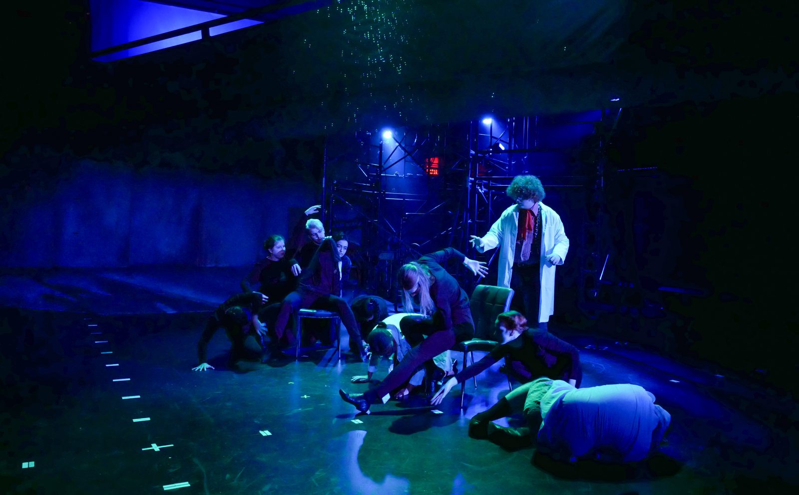 A dark stage with blue spotlights focused on 10 students at the centre of the stage. Two students sit in front with industrial scaffolding behind them.