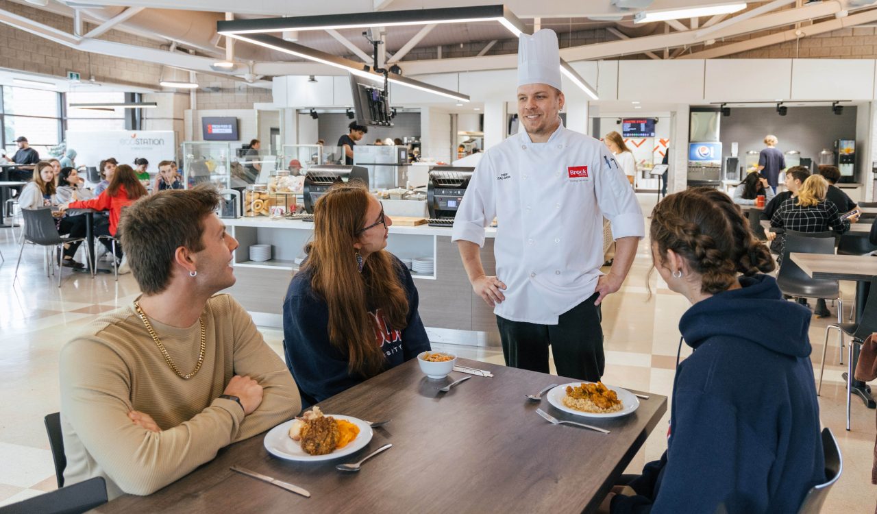 Three people sitting at a dining hall table speak with a chef who is standing at the end of the table in his white coat and chef's hat.