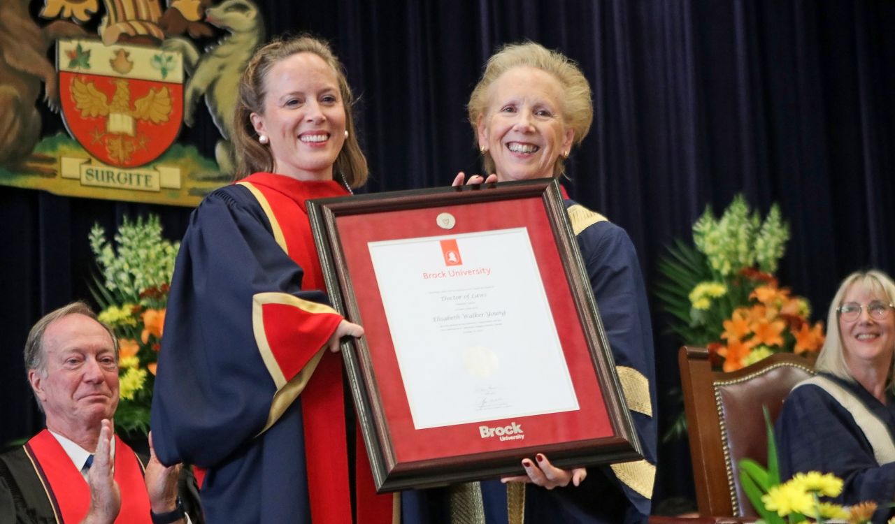 Two women in blue and red robes stand holding a framed white degree between them.