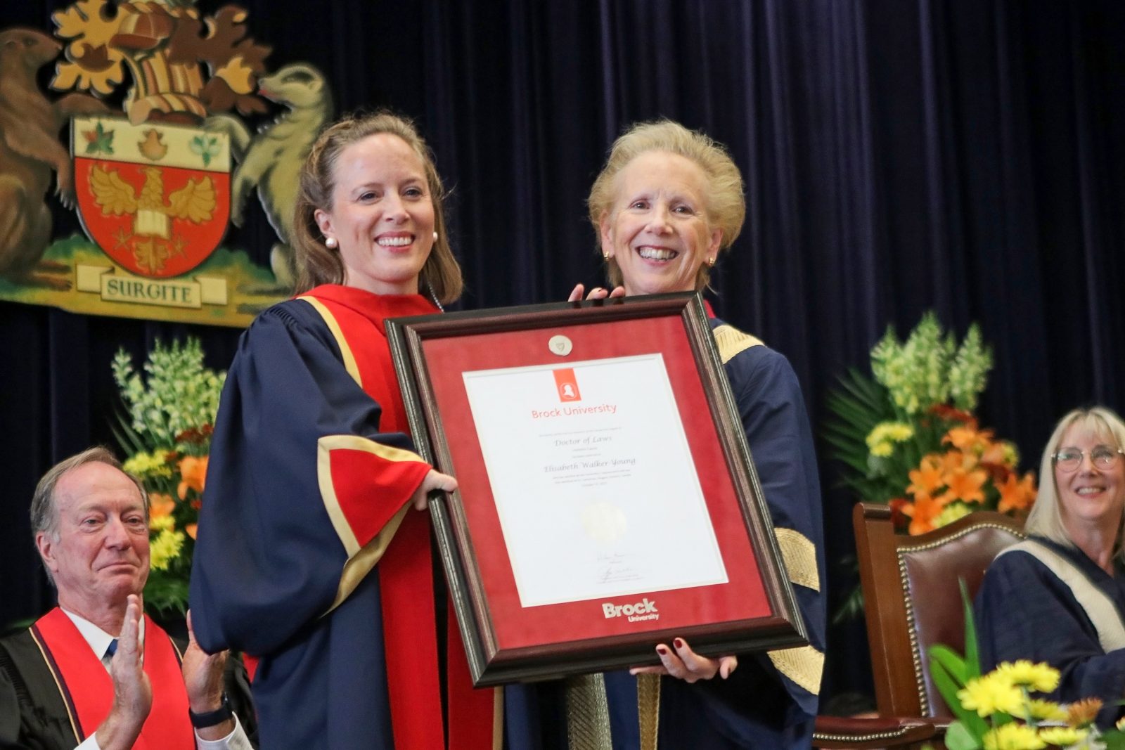 Two women in blue and red robes stand holding a framed white degree between them.