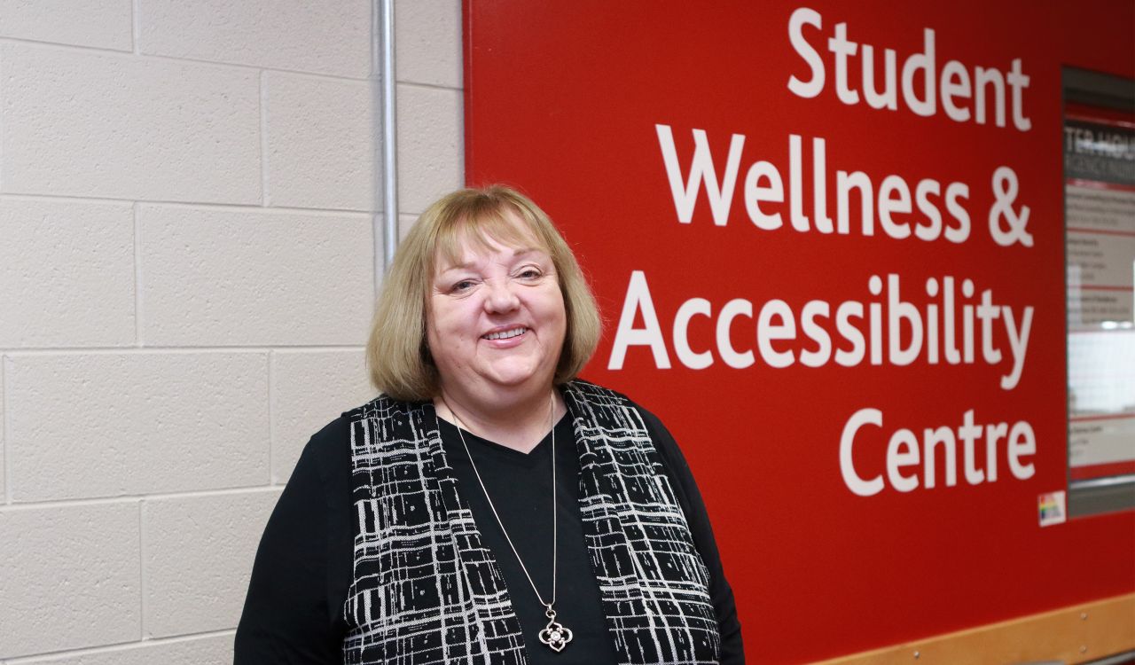 Dianne Jansen stands in front of a red wall with large white letters that read ‘Student Wellness and Accessibility Centre.’