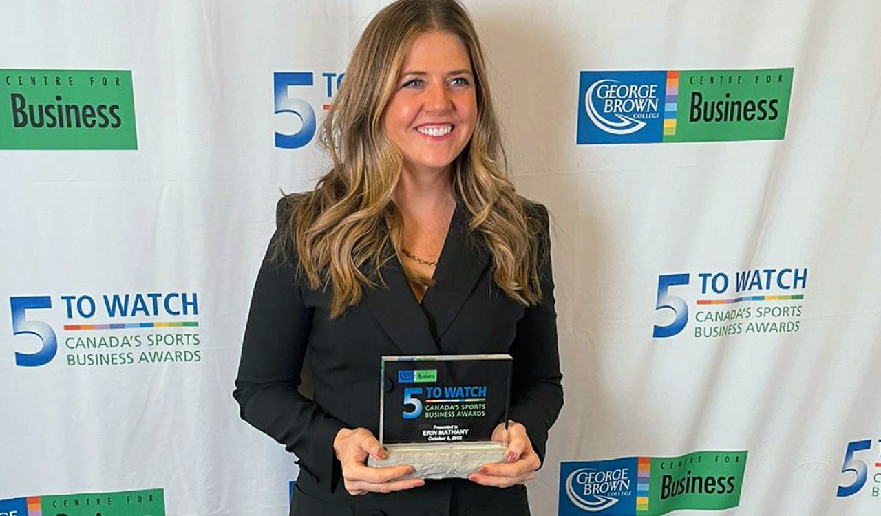 Brock University Sport Management alumna Erin Mathany holds a glass ‘5 to Watch’ award while standing in front of a backdrop filled with two repeating logos, ‘5 to Watch – Canada's Sports Business Awards’ and ‘George Brown College Centre for Business.’