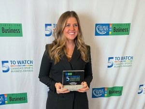 Brock University Sport Management alumna Erin Mathany holds a glass ‘5 to Watch’ award while standing in front of a backdrop filled with two repeating logos, ‘5 to Watch – Canada's Sports Business Awards’ and ‘George Brown College Centre for Business.’