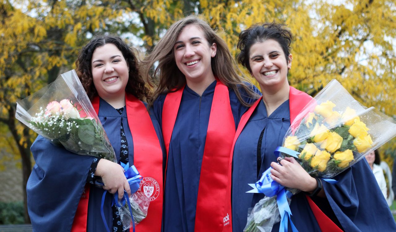 Three women in graduation gowns stand together, two of them holding bouquets of flowers.