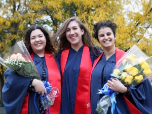 Three women in graduation gowns stand together, two of them holding bouquets of flowers.