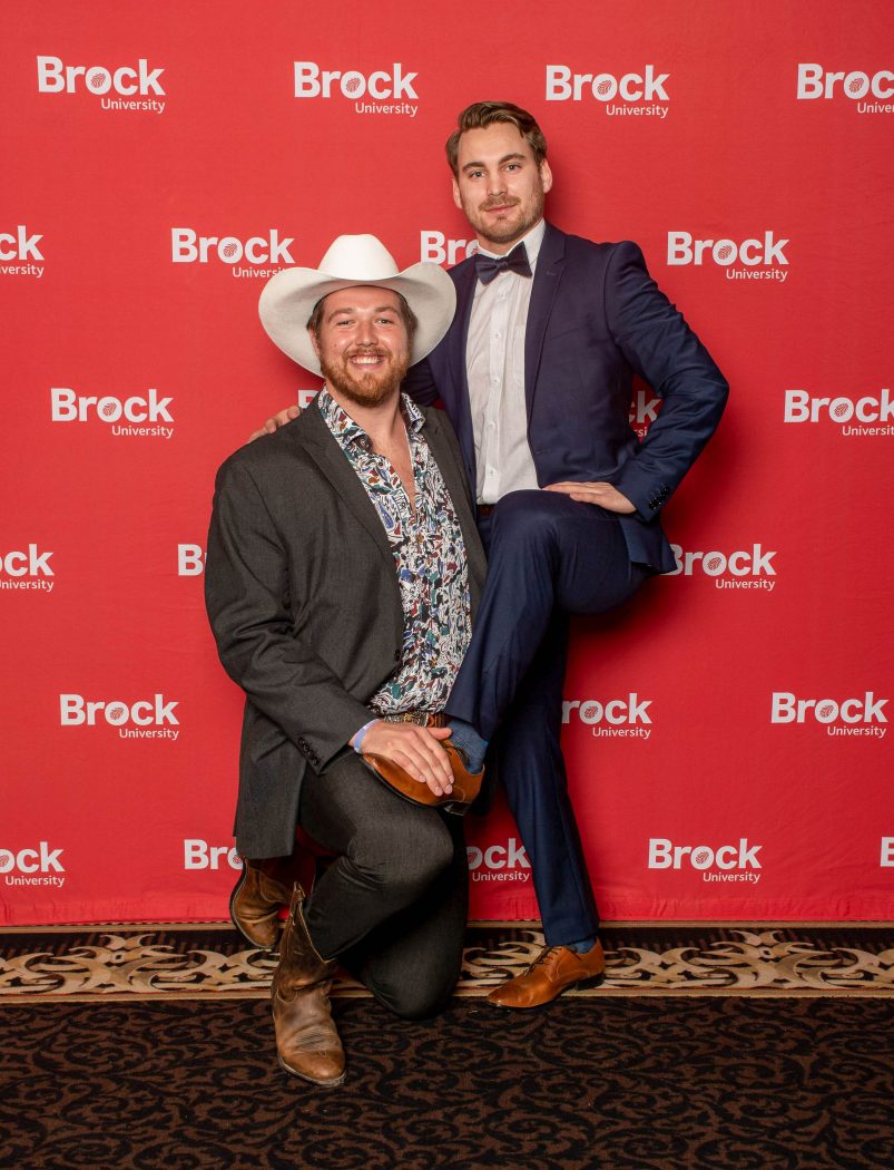 Two men pose for a photo in front of a red backdrop.