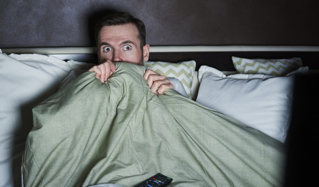 A man sitting up in bed peeks over a blanket with a remote control on his lap.