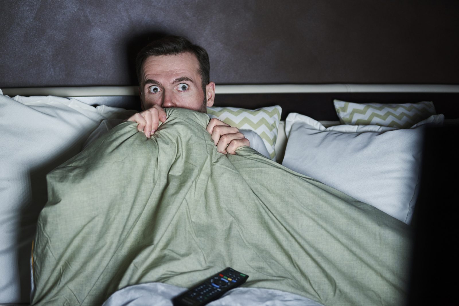 A man sitting up in bed peeks over a blanket with a remote control on his lap.