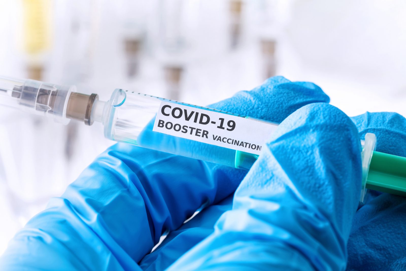 Close-up of a blue-gloved hand holding a needle labelled “COVID-19 Booster Vaccination.”