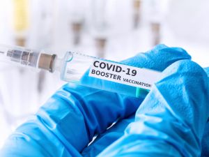 Close-up of a blue-gloved hand holding a needle labelled “COVID-19 Booster Vaccination.”