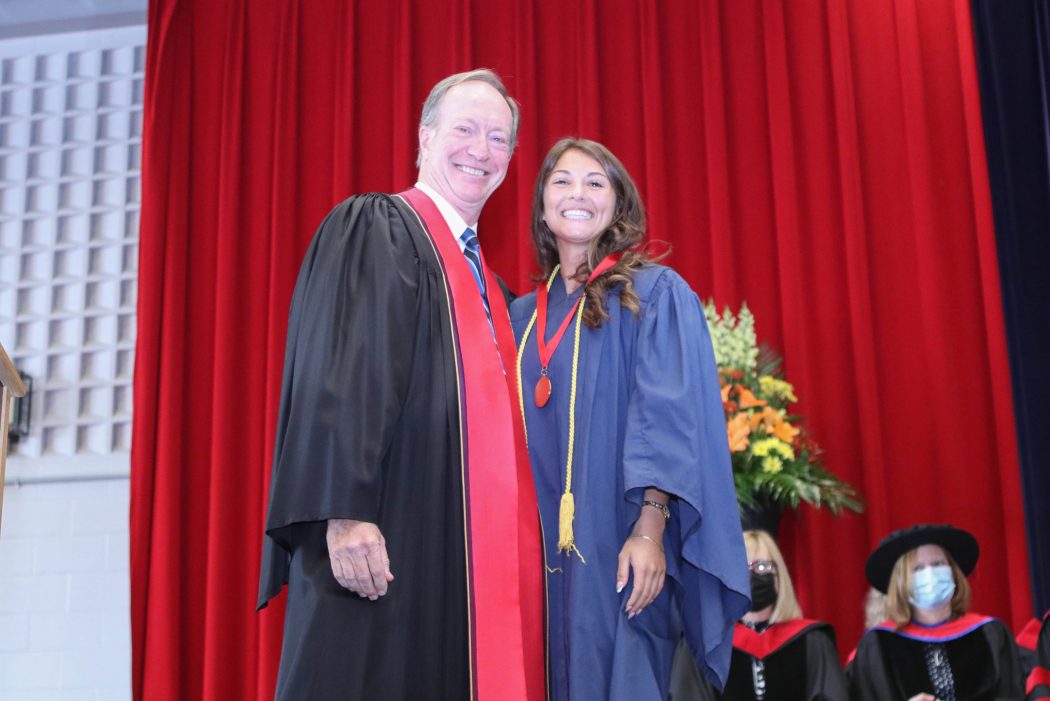 A woman in a blue robe and a red medallion stands next to a man in a black and red robe in front of a red curtain.