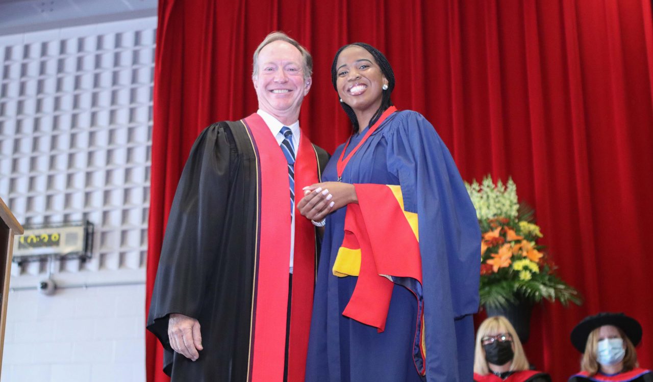 A woman in a blue robe wearing a red medallion stands next to a man in a black and red robe in front of a red curtain.