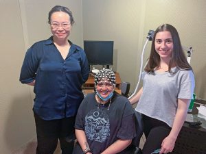 Three students pose in a laboratory with computer screen, one seated and wearing electroencephalogram net.