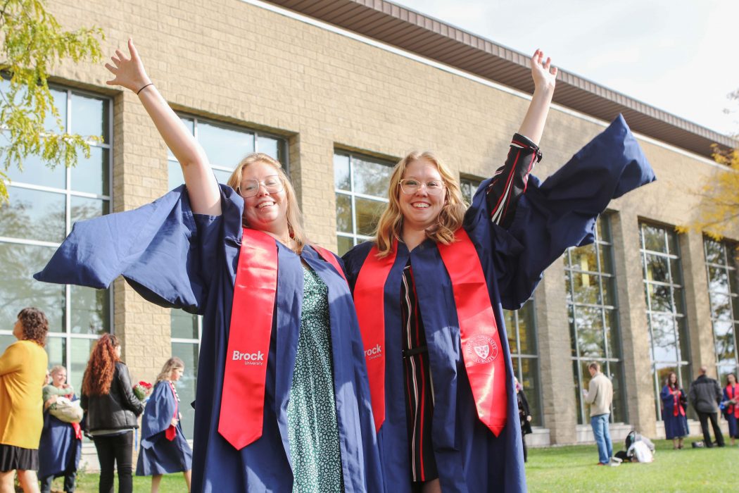 Two women in blue graduation gowns throw an arm up in celebration.