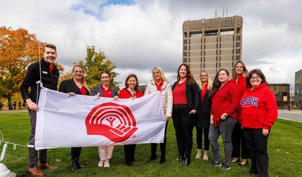 Ten people stand side by side holding a large United Way flag. Brock University's Schmon Tower stands tall in the background.