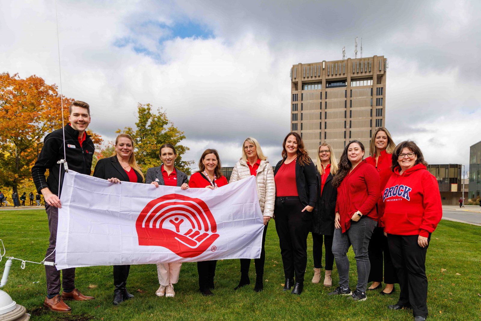 Ten people stand side by side holding a large United Way flag. Brock University's Schmon Tower stands tall in the background.