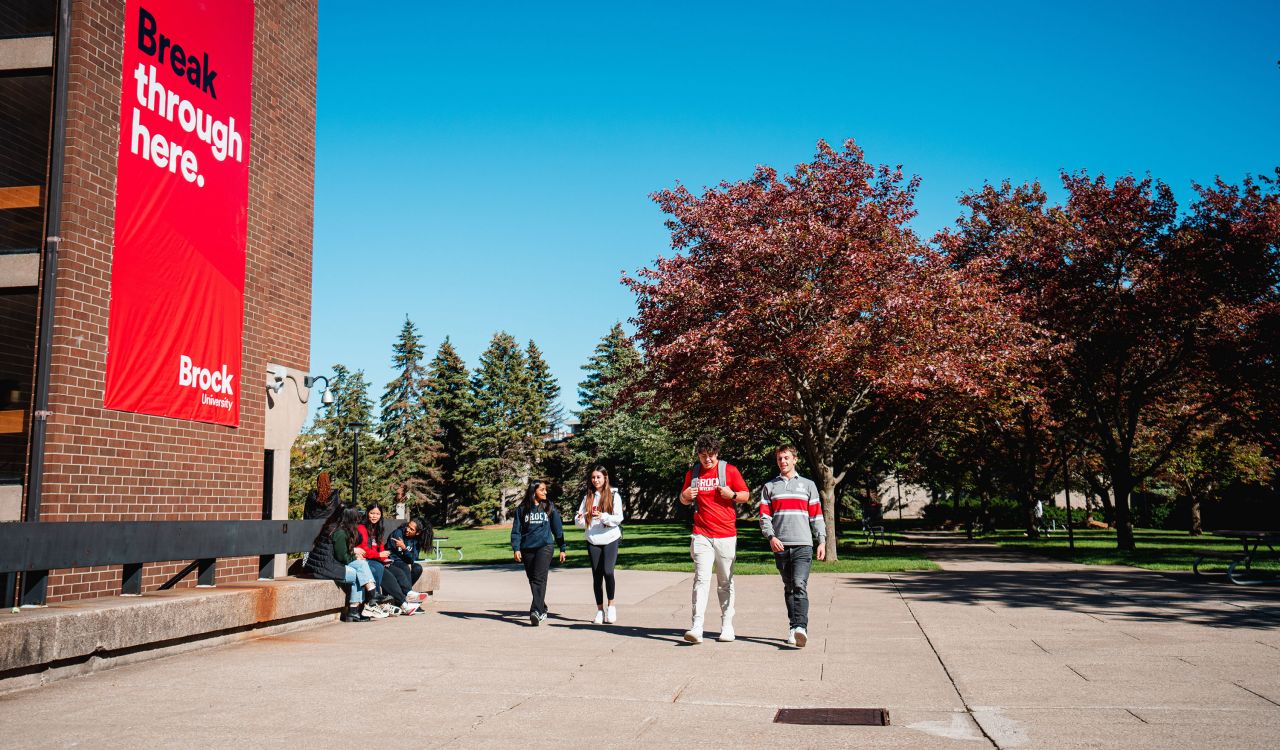 A group of students walk outside on a university campus with trees in the background and a large red banner on a brick building that reads 'Break through here.'