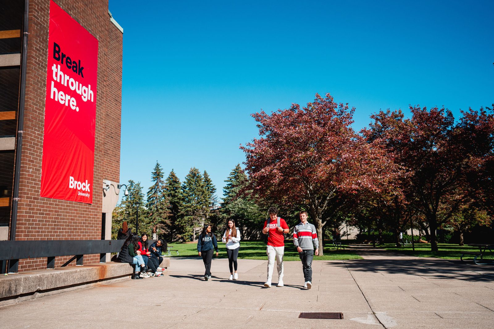 A group of students walk outside on a university campus with trees in the background and a large red banner on a brick building that reads 'Break through here.'
