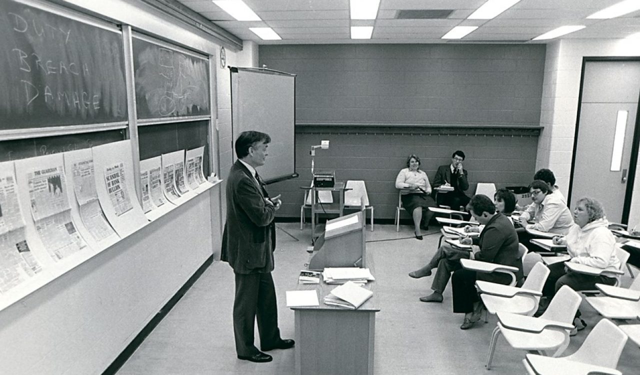In this black and white photo from 1986, Brock University Professor William “Bill”l Hull stands at the front of a classroom. Behind him is a blackboard lined with newspaper clippings. Several students sit in chairs and take notes.