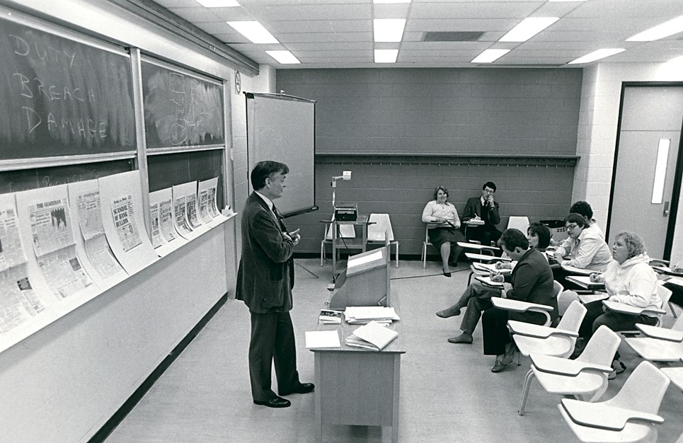 In this black and white photo from 1986, Brock University Professor William “Bill”l Hull stands at the front of a classroom. Behind him is a blackboard lined with newspaper clippings. Several students sit in chairs and take notes.