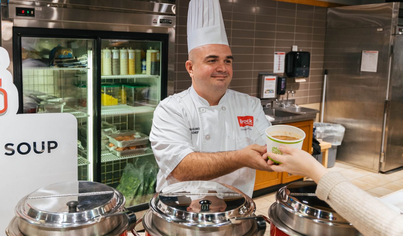 Nicholas Paulino, Executive Chef with Brock Dining Services serves a Brock University employee soup in Market Eatery. Several red cauldrons filled with soup can be seen in the foreground, while a fridge with a variety of ingredients can be seen in the background.