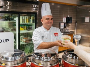 Nicholas Paulino, Executive Chef with Brock Dining Services serves a Brock University employee soup in Market Eatery. Several red cauldrons filled with soup can be seen in the foreground, while a fridge with a variety of ingredients can be seen in the background.