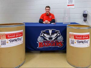 A man sits at a table with a Brock Badgers logo and two food drive bins placed in front.