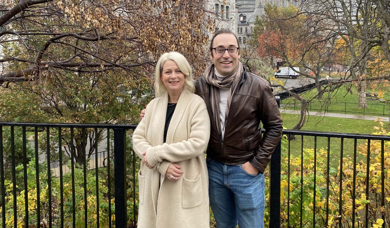 A woman in a beige jacket stands next to a man in a brown leather jacket in front of a leafy outdoor setting.