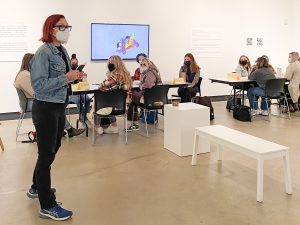 A female to the left interacting with a collective of students behind her in an art gallery.