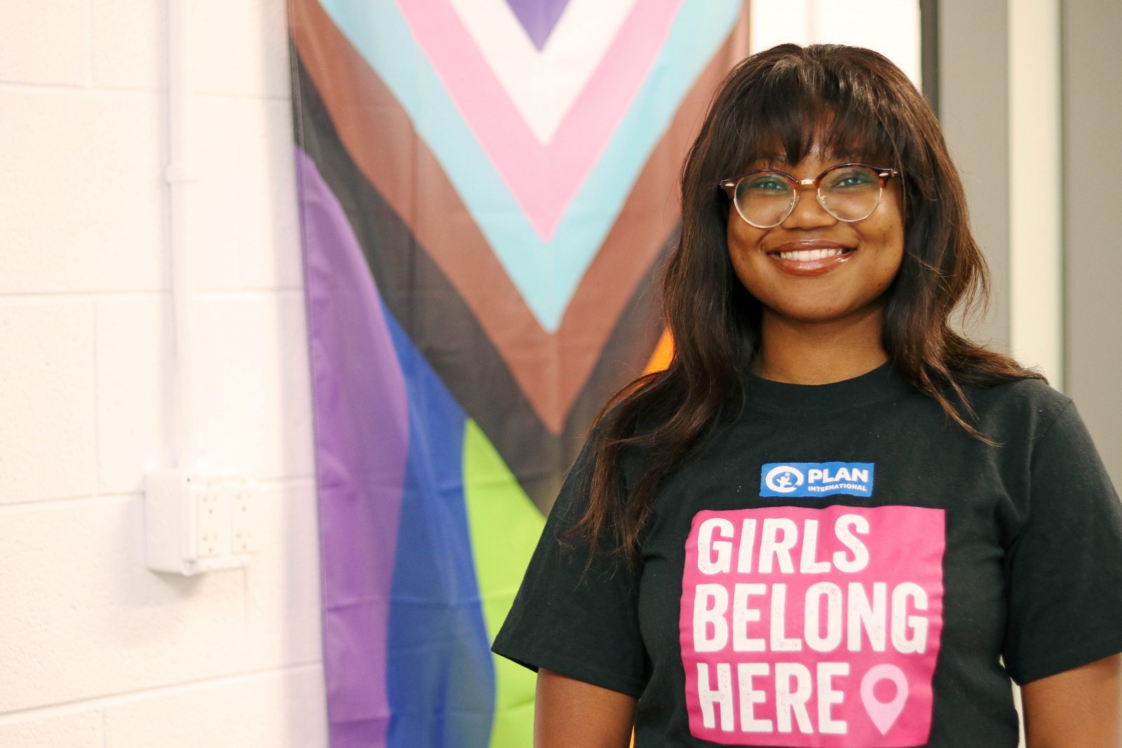 Eve Nyambiya wears a shirt that reads 'Girls Belong Here'. She is standing in front of the modern Pride flag, which includes stripes to represent the experiences of People of Colour, as well as stripes to represent people who identify as transgender, gender nonconforming and/or undefined.