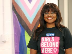 Eve Nyambiya wears a shirt that reads 'Girls Belong Here'. She is standing in front of the modern Pride flag, which includes stripes to represent the experiences of People of Colour, as well as stripes to represent people who identify as transgender, gender nonconforming and/or undefined.