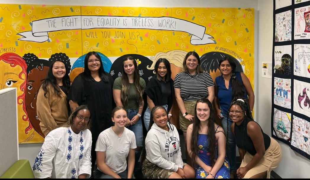 Eleven young women, six standing in a row and five kneeling in a second row, pose for a photo in front of a large mural with painted images of women’s faces on a yellow background with text that reads “The fight for equity is tireless work. Will you join us?” 