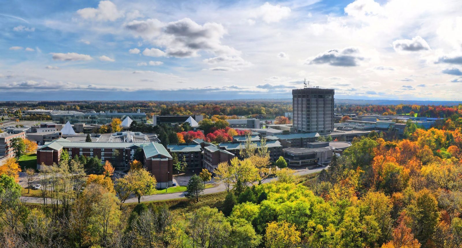 An aerial photograph of Brock University. A campus filled with buildings is surrounded by greenspace. Some of the trees' leaves have turned red, orange and yellow for fall. In the distance, Lake Ontario glistens under a blue sky peppered with fluffy white clouds.