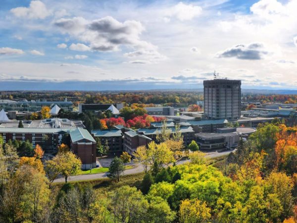 An aerial photograph of Brock University. A campus filled with buildings is surrounded by greenspace. Some of the trees' leaves have turned red, orange and yellow for fall. In the distance, Lake Ontario glistens under a blue sky peppered with fluffy white clouds.