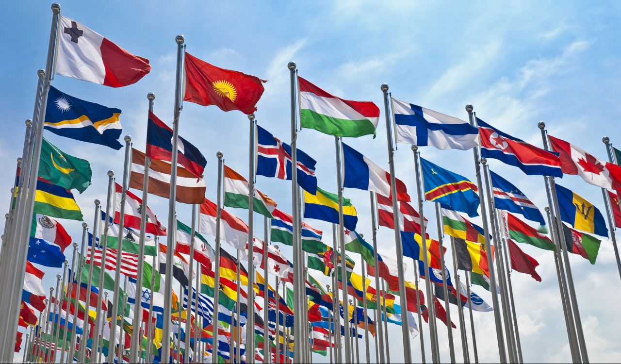 Various flags from around the world fly on poles against a blue sky background.