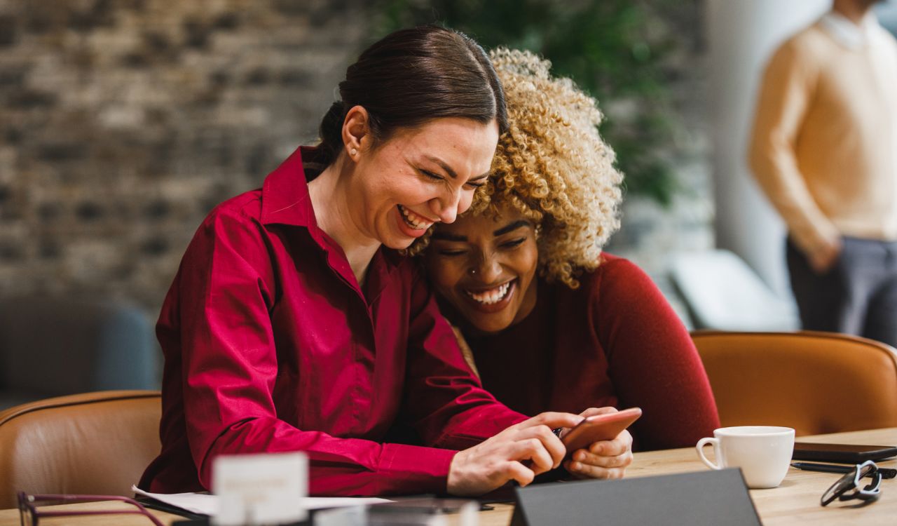 Two women seated in a coffee shop laugh at an image on a smartphone