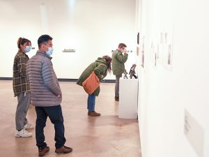 Four individuals in an open art gallery examining photographs put up on white walls.